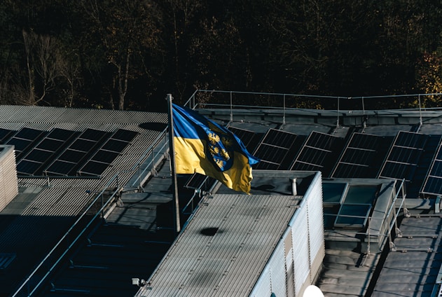 A flag with yellow and blue colors featuring a coat of arms is prominently displayed on a rooftop equipped with solar panels. The rooftop is surrounded by trees, indicating a mix of natural and industrial elements.