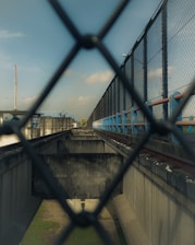 A view through a chain-link fence, focusing on an industrial setting. The image contains a corridor-like structure with high concrete walls and metal railings, extending towards the horizon. There is a pathway or track on one side, bordered by blue and red elements. The sky is visible above, with some clouds.