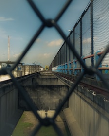 A view through a chain-link fence, focusing on an industrial setting. The image contains a corridor-like structure with high concrete walls and metal railings, extending towards the horizon. There is a pathway or track on one side, bordered by blue and red elements. The sky is visible above, with some clouds.
