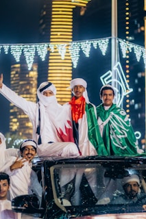 A group of men in traditional attire are standing on top of a vehicle. They are holding flags and celebrating, likely during a national event. Bright city lights and skyscrapers are visible in the background, creating a festive and vibrant atmosphere with illuminated decorations.