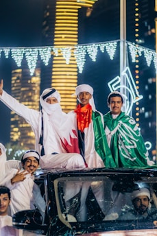 A group of men in traditional attire are standing on top of a vehicle. They are holding flags and celebrating, likely during a national event. Bright city lights and skyscrapers are visible in the background, creating a festive and vibrant atmosphere with illuminated decorations.