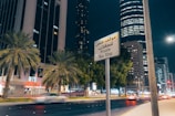 A nighttime cityscape featuring tall buildings, palm trees, and a busy street with moving cars. A sign in both Arabic and English reads 'Private Bus Stop.' The buildings are illuminated, and the street is well-lit with a modern urban ambiance.