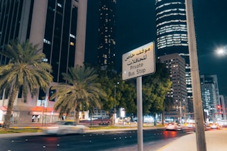 A nighttime cityscape featuring tall buildings, palm trees, and a busy street with moving cars. A sign in both Arabic and English reads 'Private Bus Stop.' The buildings are illuminated, and the street is well-lit with a modern urban ambiance.