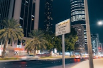 A nighttime cityscape featuring tall buildings, palm trees, and a busy street with moving cars. A sign in both Arabic and English reads 'Private Bus Stop.' The buildings are illuminated, and the street is well-lit with a modern urban ambiance.