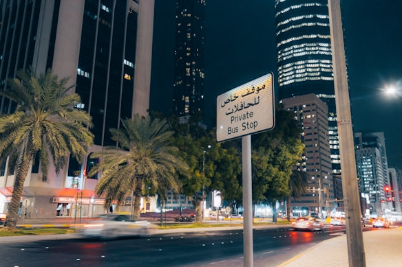 A nighttime cityscape featuring tall buildings, palm trees, and a busy street with moving cars. A sign in both Arabic and English reads 'Private Bus Stop.' The buildings are illuminated, and the street is well-lit with a modern urban ambiance.
