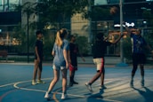 A group of young people are playing basketball on an outdoor court during the evening. One player is jumping to shoot the ball, while others are either preparing to rebound or observing. The court is surrounded by a high fence and there are trees and buildings visible in the background, adding an urban atmosphere.