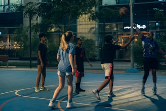 A group of young people are playing basketball on an outdoor court during the evening. One player is jumping to shoot the ball, while others are either preparing to rebound or observing. The court is surrounded by a high fence and there are trees and buildings visible in the background, adding an urban atmosphere.