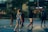 A group of young people are playing basketball on an outdoor court during the evening. One player is jumping to shoot the ball, while others are either preparing to rebound or observing. The court is surrounded by a high fence and there are trees and buildings visible in the background, adding an urban atmosphere.