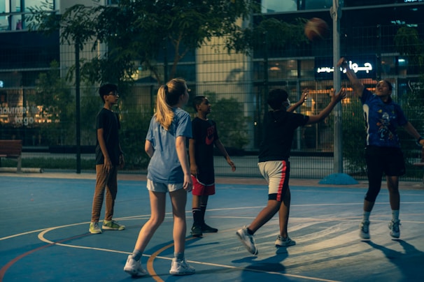 A group of young people are playing basketball on an outdoor court during the evening. One player is jumping to shoot the ball, while others are either preparing to rebound or observing. The court is surrounded by a high fence and there are trees and buildings visible in the background, adding an urban atmosphere.