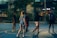 A group of young people are playing basketball on an outdoor court during the evening. One player is jumping to shoot the ball, while others are either preparing to rebound or observing. The court is surrounded by a high fence and there are trees and buildings visible in the background, adding an urban atmosphere.