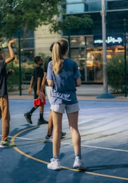 A group of young people are playing basketball on an outdoor court. The scene is set in the evening with artificial lighting illuminating the area. They are casually dressed in sports attire, and there is a building with a neon sign visible in the background.