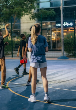 A group of young people are playing basketball on an outdoor court. The scene is set in the evening with artificial lighting illuminating the area. They are casually dressed in sports attire, and there is a building with a neon sign visible in the background.
