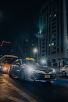 A silver taxi is driving through a city street at night, with its headlights shining brightly. Tall buildings with illuminated windows tower in the background. Other vehicles, including a bus, are also present on the wet road, reflecting the city lights.