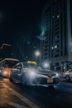 A silver taxi is driving through a city street at night, with its headlights shining brightly. Tall buildings with illuminated windows tower in the background. Other vehicles, including a bus, are also present on the wet road, reflecting the city lights.
