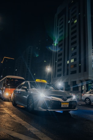 A silver taxi is driving through a city street at night, with its headlights shining brightly. Tall buildings with illuminated windows tower in the background. Other vehicles, including a bus, are also present on the wet road, reflecting the city lights.