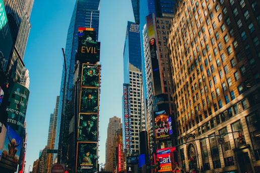 Tall buildings adorned with colorful advertisements and digital screens, bathed in warm sunlight. The iconic urban setting features bustling skyscrapers and vibrant displays.