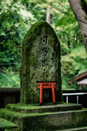 A moss-covered stone tablet featuring engraved Japanese characters stands in a lush, green forest setting. In front of the stone is a small, vibrant red torii gate, adding contrast to the natural surroundings. The scene is tranquil, with a sense of historical or cultural significance.