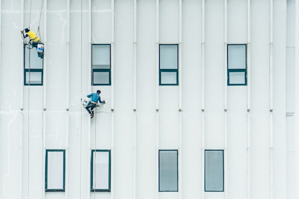 Two workers are suspended on ropes, painting or cleaning the exterior of a tall building. They are positioned on a vertical wall with multiple rectangular windows, dressed in safety gear.
