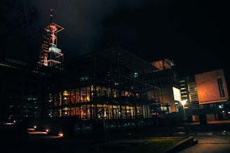 A modern building structure illuminated at night, featuring glass walls and bright interior lighting. The architecture includes metal frameworks and angular designs, creating a futuristic appearance. A tall illuminated spire extends from the top of the building, adding an element of industrial design. Dim streetlights and minimal outdoor lighting emphasize the darkness surrounding the structure.