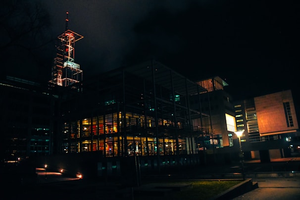 A modern building structure illuminated at night, featuring glass walls and bright interior lighting. The architecture includes metal frameworks and angular designs, creating a futuristic appearance. A tall illuminated spire extends from the top of the building, adding an element of industrial design. Dim streetlights and minimal outdoor lighting emphasize the darkness surrounding the structure.