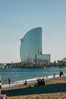 A modern, sail-shaped skyscraper stands adjacent to a sandy beach filled with people enjoying the sun. The foreground shows groups of people sitting and walking on the beach, while some are in the water. A clear blue sky with a seagull flying by adds to the serene atmosphere.