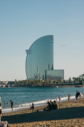 A modern, sail-shaped skyscraper stands adjacent to a sandy beach filled with people enjoying the sun. The foreground shows groups of people sitting and walking on the beach, while some are in the water. A clear blue sky with a seagull flying by adds to the serene atmosphere.