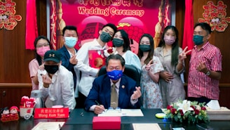 A group of people gathered in an indoor setting, wearing masks and displaying happy expressions. In the center, a man in a suit is seated at a desk, signing a document. The backdrop includes a festive sign reading 'Wedding Ceremony' with decorative elements and flowers around.