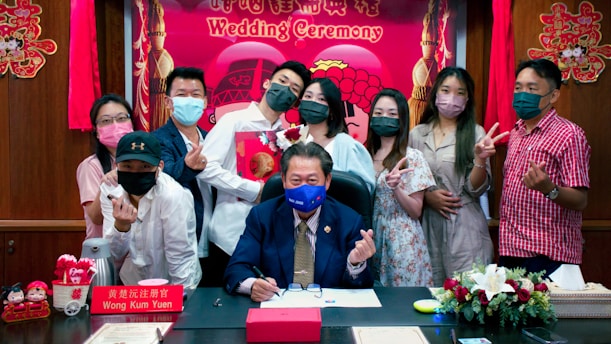 A group of people gathered in an indoor setting, wearing masks and displaying happy expressions. In the center, a man in a suit is seated at a desk, signing a document. The backdrop includes a festive sign reading 'Wedding Ceremony' with decorative elements and flowers around.