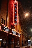 A brightly lit marquee of the Mayan theater displays several movie titles, framed by neon lights. Snow gently falls in the foreground, adding a wintry atmosphere to the urban night scene. The theater sign is prominently lit, with the building's detailed facade visible.