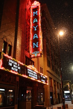 A brightly lit marquee of the Mayan theater displays several movie titles, framed by neon lights. Snow gently falls in the foreground, adding a wintry atmosphere to the urban night scene. The theater sign is prominently lit, with the building's detailed facade visible.