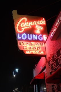 A neon sign glows brightly against the night sky, displaying the text 'Gennaro's Lounge' with a martini glass icon, followed by 'Italian Food Pizza'. The sign is attached to the side of a building, which has red awnings. A street lamp is visible below, casting a soft light.