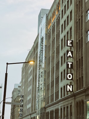 Tall buildings with large signage displaying the word 'EATON', featuring a mix of classical and modern architectural styles. Illuminated streetlamp and a signpost are visible in the foreground.