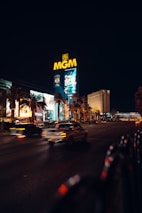 A nighttime cityscape featuring the brightly lit MGM sign dominating the scene. The foreground shows blurred motion of cars on a wide road, flanked by illuminated billboards and palm trees. The sky is dark, contrasting with the vibrant lights of the city.