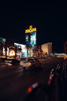 A nighttime cityscape featuring the brightly lit MGM sign dominating the scene. The foreground shows blurred motion of cars on a wide road, flanked by illuminated billboards and palm trees. The sky is dark, contrasting with the vibrant lights of the city.