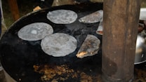 Flatbreads are cooking on a large round griddle. Several pieces are laid out and one is partially filled and folded. A metal kettle is visible in the background next to a tall cylindrical metal object.