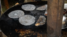 Flatbreads are cooking on a large round griddle. Several pieces are laid out and one is partially filled and folded. A metal kettle is visible in the background next to a tall cylindrical metal object.