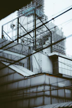 A construction site with high-rise buildings in the background and a complex framework of metal scaffolding in the forefront. Multiple wires or cables are crisscrossing in the scene, adding to the visual complexity. The structures appear to be under construction or renovation.