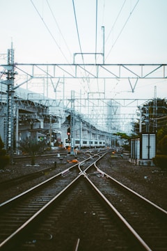 Railway tracks leading into the distance, with overhead power lines and industrial structures nearby. A train is visible in the background, and there is greenery on the right side. The tracks interconnect, indicating a complex rail junction.