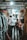 A narrow market alley with several individuals looking at and selecting fabrics. Various types of cloth are displayed on the left side, hanging from racks. The passage is dimly lit with a bright light overhead. People appear to be shopping, casually browsing the merchandise.