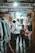 A narrow market alley with several individuals looking at and selecting fabrics. Various types of cloth are displayed on the left side, hanging from racks. The passage is dimly lit with a bright light overhead. People appear to be shopping, casually browsing the merchandise.