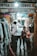 A narrow market alley with several individuals looking at and selecting fabrics. Various types of cloth are displayed on the left side, hanging from racks. The passage is dimly lit with a bright light overhead. People appear to be shopping, casually browsing the merchandise.