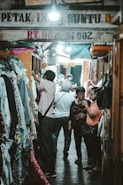 A narrow market alley with several individuals looking at and selecting fabrics. Various types of cloth are displayed on the left side, hanging from racks. The passage is dimly lit with a bright light overhead. People appear to be shopping, casually browsing the merchandise.