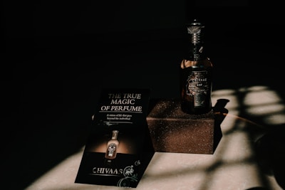 A bottle of perfume labeled 'CHIVAAS' sits on a textured rectangular block, accompanied by a promotional piece of paper with the phrase 'The True Magic of Perfume'. The scene is bathed in dramatic lighting creating strong shadows on the surface.
