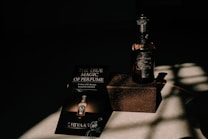 A bottle of perfume labeled 'CHIVAAS' sits on a textured rectangular block, accompanied by a promotional piece of paper with the phrase 'The True Magic of Perfume'. The scene is bathed in dramatic lighting creating strong shadows on the surface.