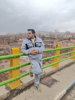 A person in a grey tracksuit is leaning against a bridge railing with a thoughtful expression. The bridge railings are painted in green and yellow. The background features a rural landscape with leafless trees, a few scattered houses, and distant mountains under a cloudy sky.