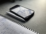 A notebook with handwritten notes in blue ink lies open on a dark tabletop. Beside it is a white pen and a smartphone with a case, showing a reflection of clouds on its screen.