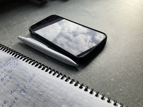 A notebook with handwritten notes in blue ink lies open on a dark tabletop. Beside it is a white pen and a smartphone with a case, showing a reflection of clouds on its screen.