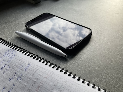 A notebook with handwritten notes in blue ink lies open on a dark tabletop. Beside it is a white pen and a smartphone with a case, showing a reflection of clouds on its screen.