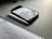 A notebook with handwritten notes in blue ink lies open on a dark tabletop. Beside it is a white pen and a smartphone with a case, showing a reflection of clouds on its screen.