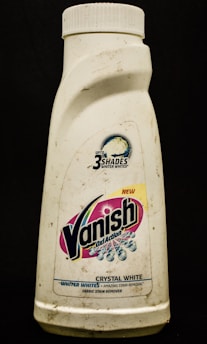 A plastic bottle labeled with the brand Vanish Oxi Action, featuring text promoting its ability to make clothes whiter. The bottle appears somewhat dirty and has a white cap.